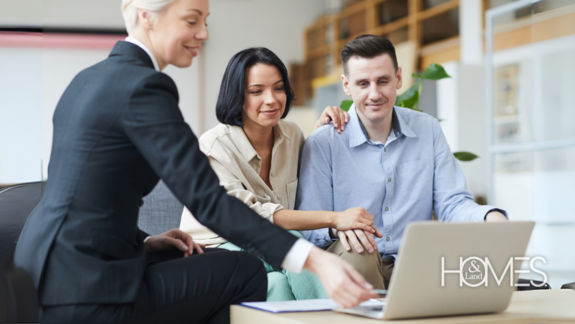 Real estate agent in a professional setting discussing property details with a couple. The agent is pointing to a laptop screen, guiding the clients through marketing or listing strategies. A bright and welcoming office environment sets a professional tone, with the Homes & Land logo subtly displayed in the corner.